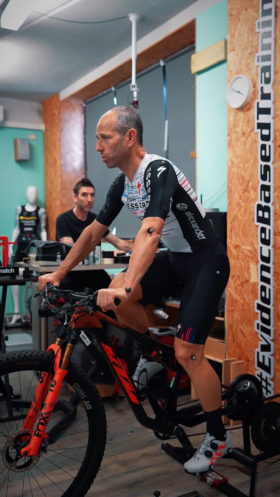 Luca Riceputi on a stationary bicycle during a STT Systems 3DMA motion analysis, with markers on his shoulders and knees to capture performance data.