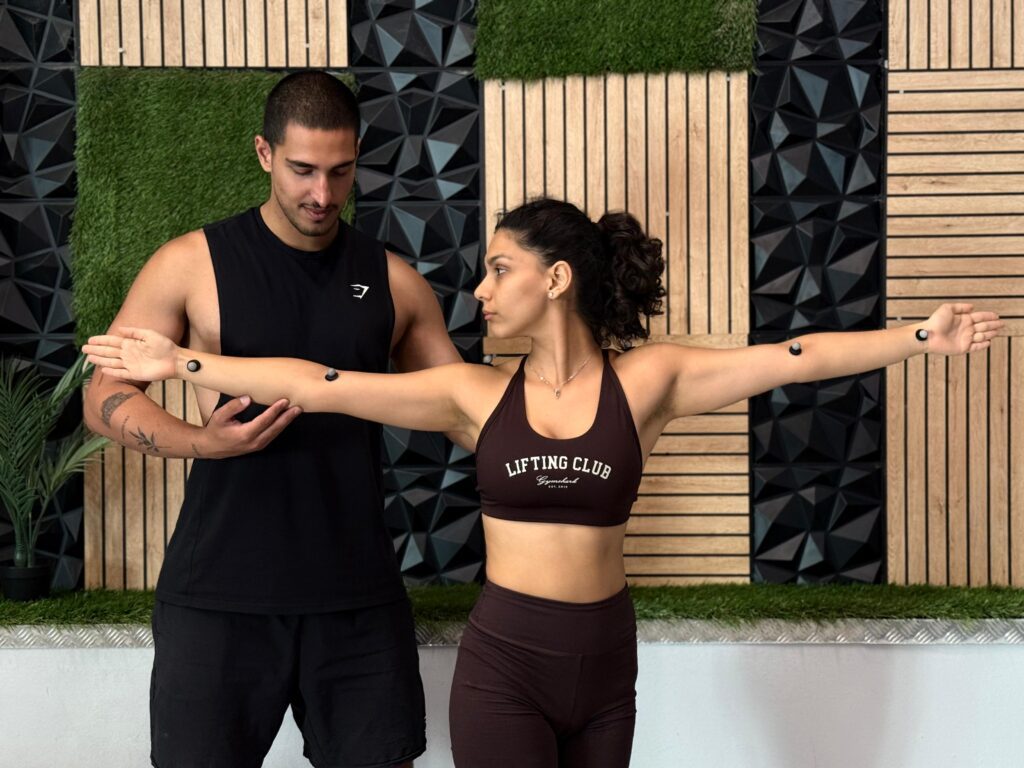 A physical therapist helping a woman with her posture, with her arms extended. She has motion markers on her arms for a movement assessment.