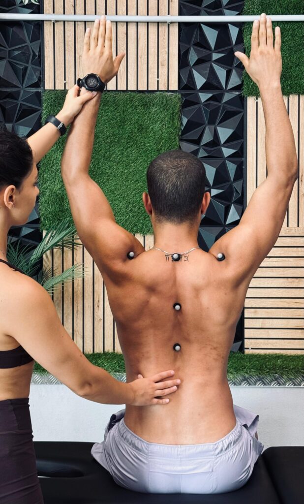 A female therapist placing a motion capture marker on a man's back. He is seated and shirtless, with markers already placed on his spine.