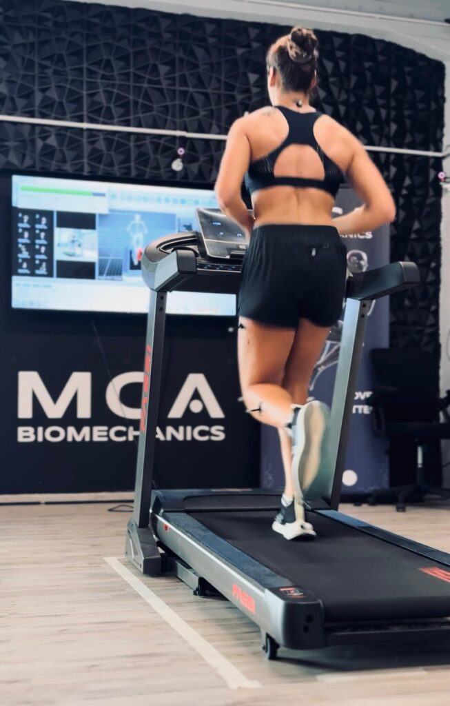 Woman running on a treadmill during a 3D biomechanical gait analysis. The image shows reflective markers on her legs and motion data on a screen in the background.