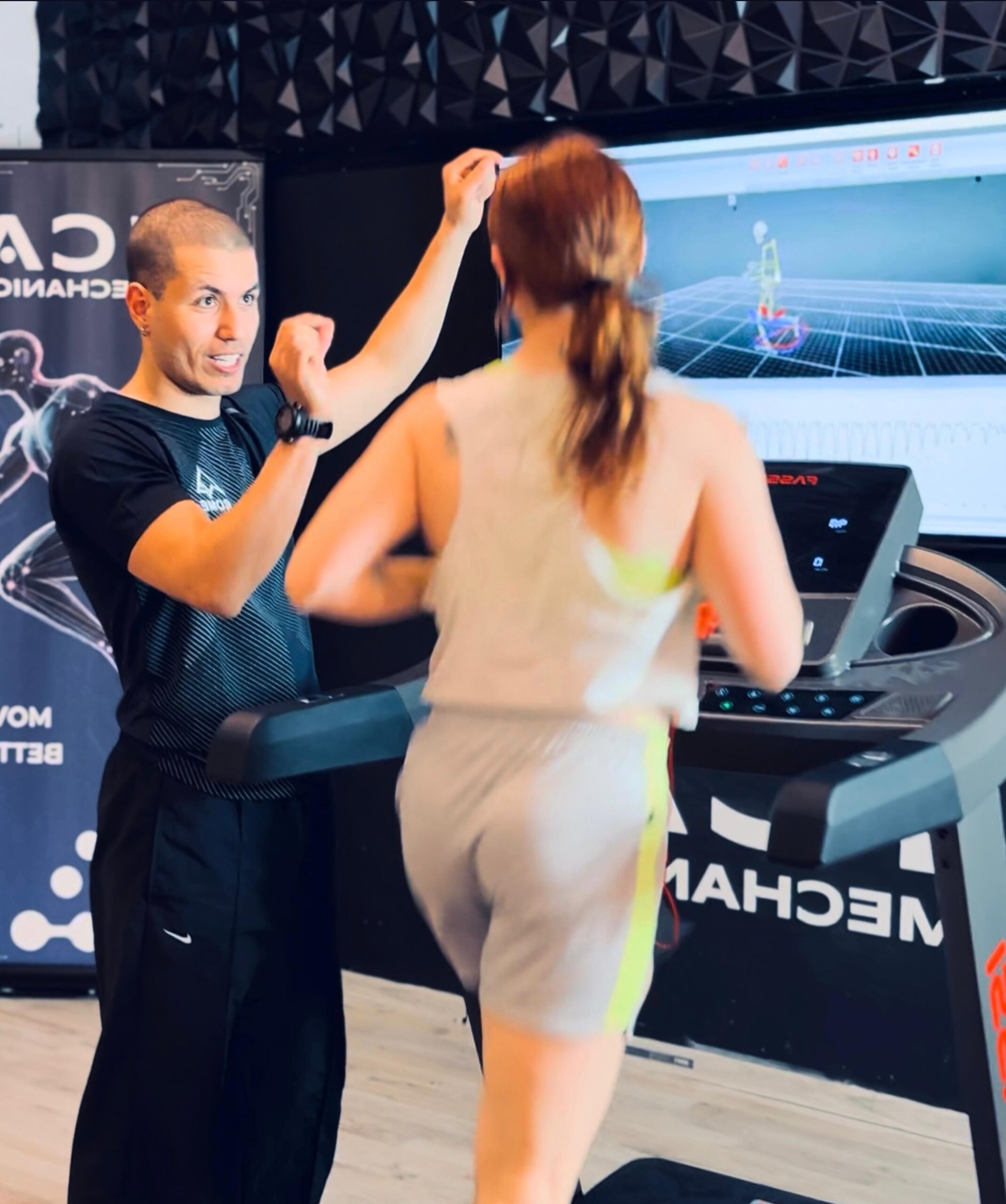 A sports performance specialist instructing and motivating a woman as she runs on a treadmill. The image shows the motion analysis data on a screen behind them.
